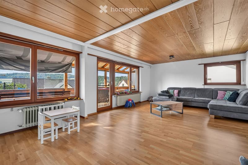 Bright living room with wooden floors, large windows, and a view of the landscape. Features a grey sectional sofa, a wooden coffee table, a small white table with chairs, and a radiator.