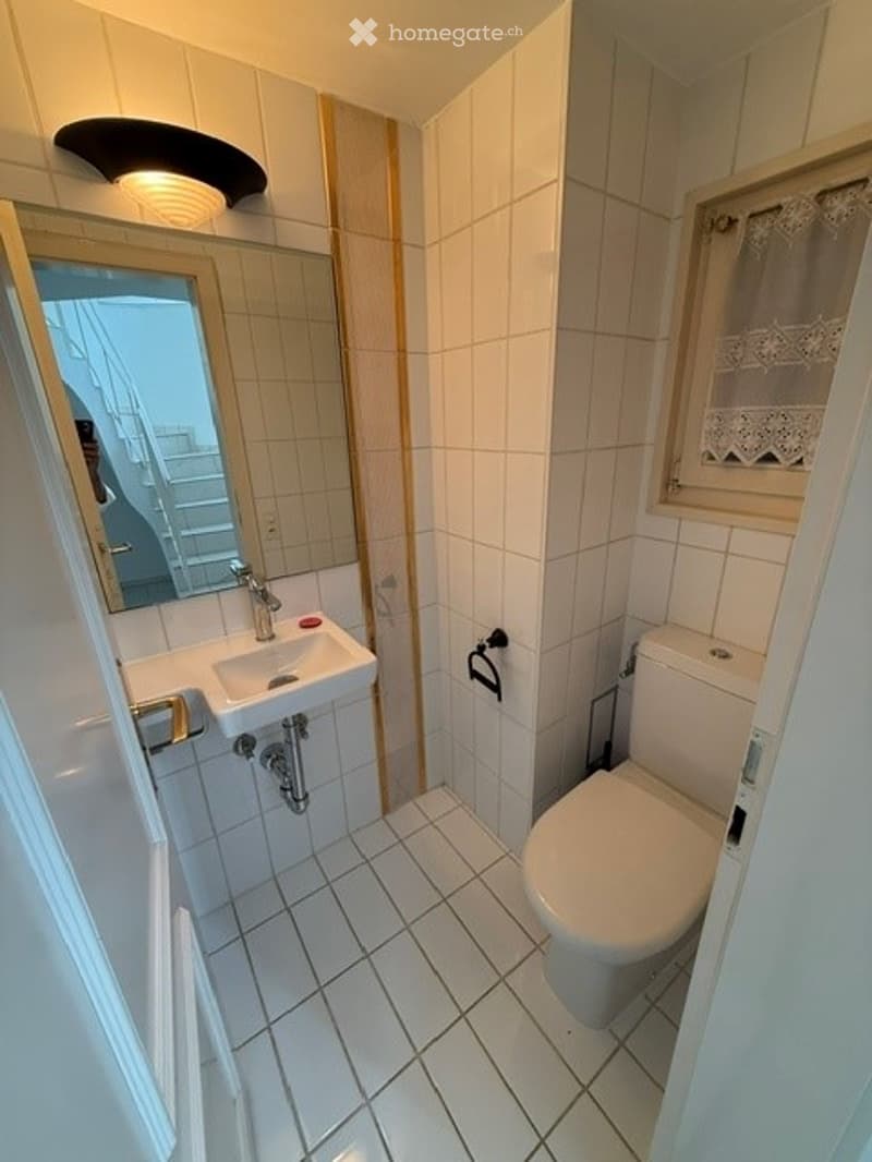Small bathroom with white tiled walls and floor, a sink with a mirror above it, and a toilet. A window with a lace curtain is visible on the right side.