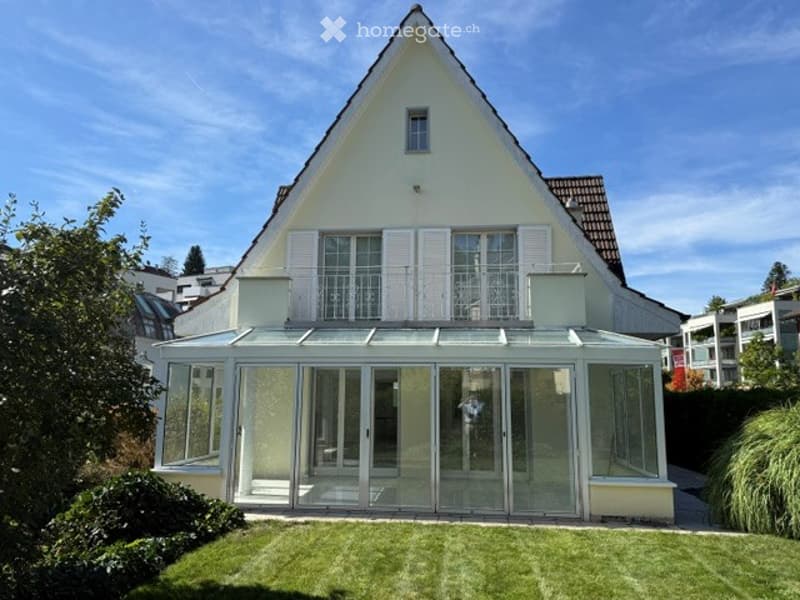 Single-family house with a modern design, featuring a large glass-enclosed patio area attached to the main structure. The house has a pitched roof and white exterior walls.