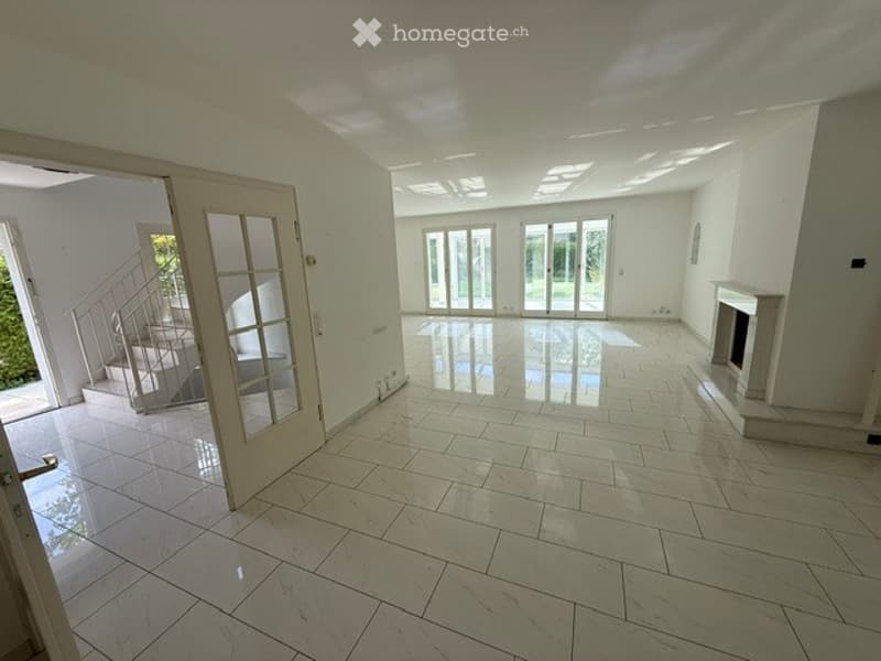 Bright, empty living space with large glass doors leading to an outdoor area, a staircase, and a fireplace. The floor is covered with large, light-colored tiles.