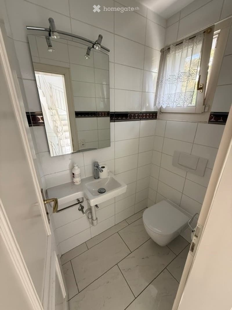 Modern bathroom with white tiles, a large mirror, a sink, a toilet, and a window with a curtain.