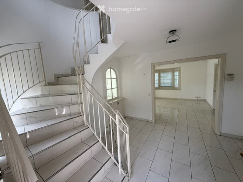 Spacious, well-lit hallway with a modern white staircase featuring a curved metal railing and a small landing. The hallway has tiled flooring and white walls, with a doorway leading to an adjacent room.