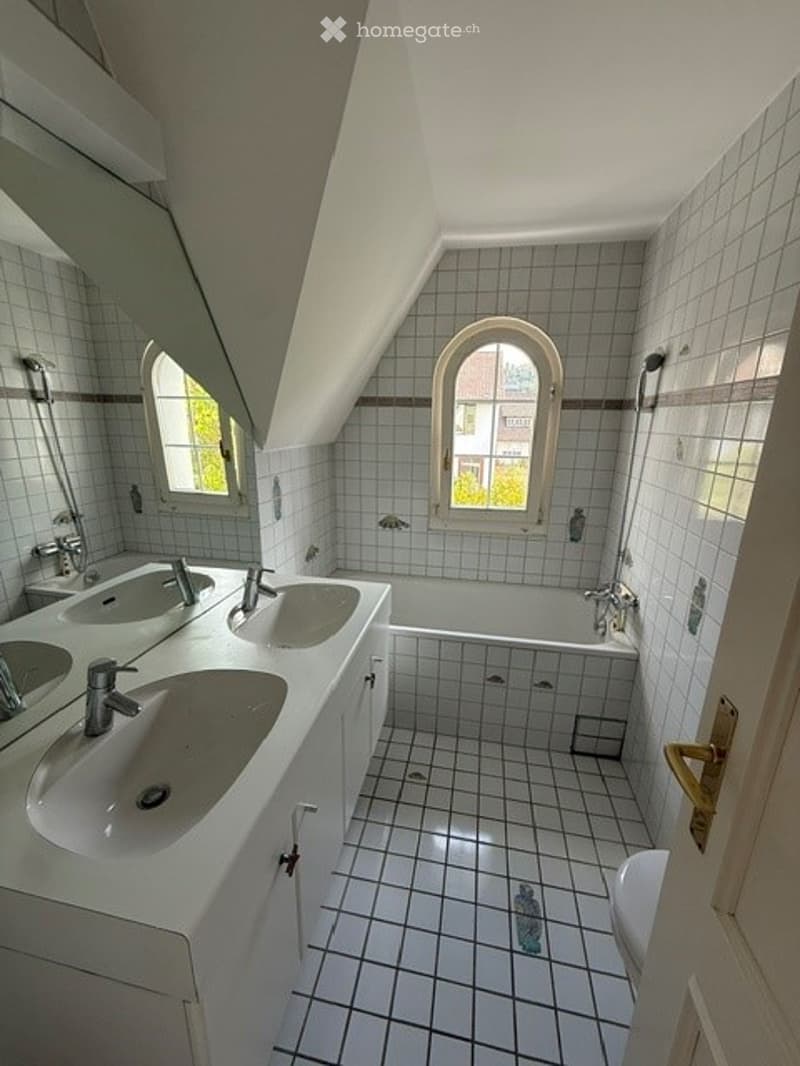 Bright bathroom with white tiled walls and floor, featuring a double sink vanity, a bathtub, and two arched windows allowing natural light.