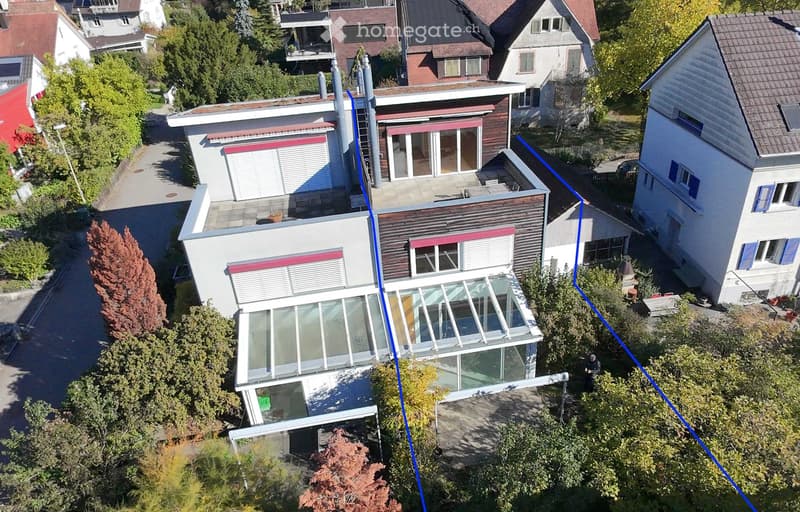 Aerial view of a modern two-story house with large glass windows and a flat roof, surrounded by trees and neighboring houses.