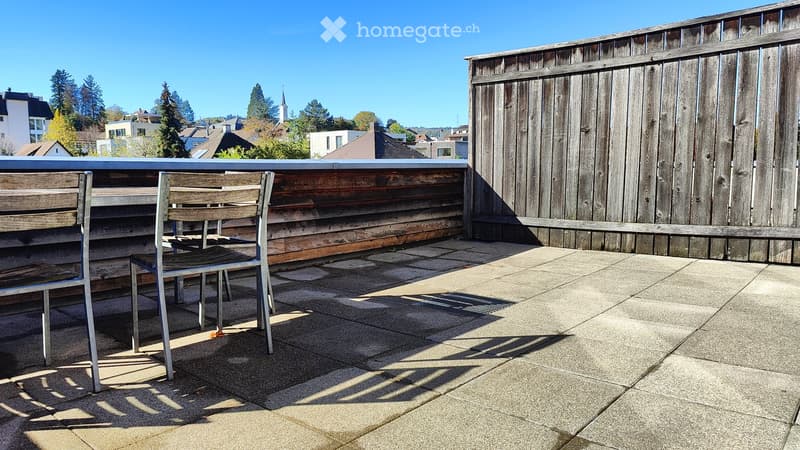 Wooden balcony with a metal railing, two chairs, and a view of the city and trees.