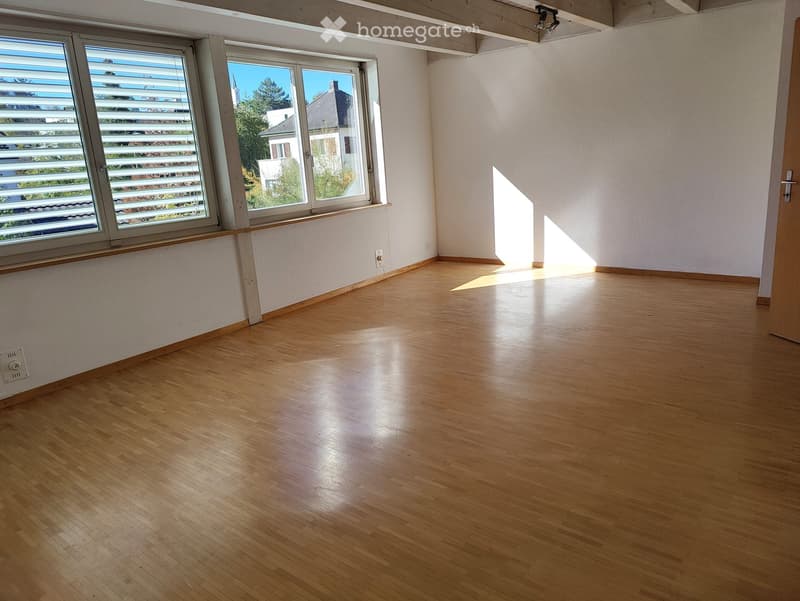 Empty room with wooden flooring, white walls, large windows with blinds, and a door leading to another area.