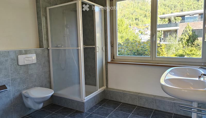 Modern bathroom with a toilet, shower, and sink. Large window offers a view of greenery outside.