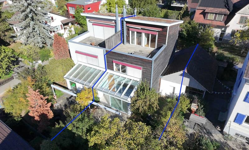 Aerial view of a modern, multi-story house with a flat roof, large windows, and a surrounding garden area.