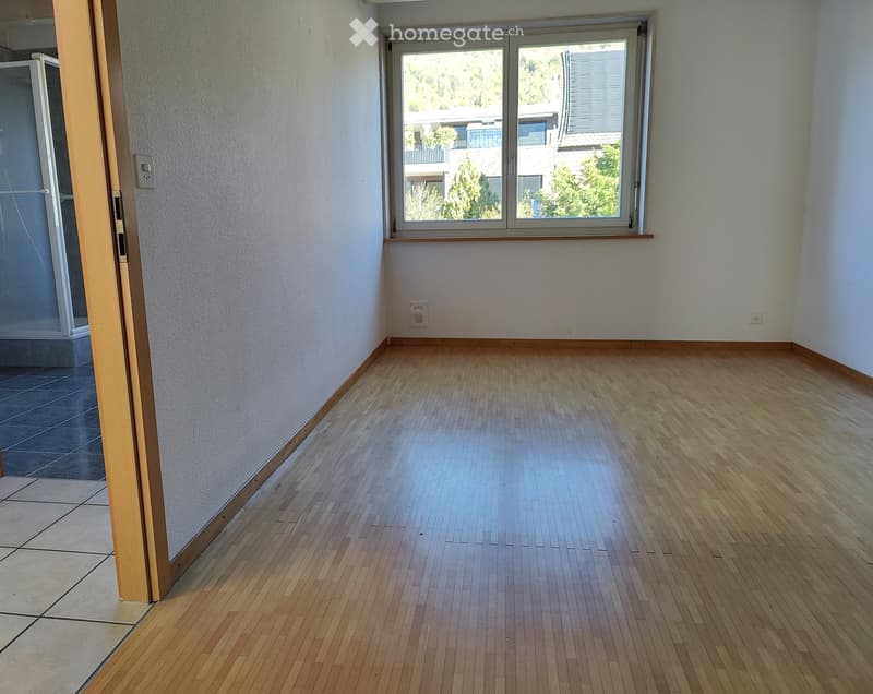 Empty room with wooden flooring, white walls, and a large window with a view of greenery outside.