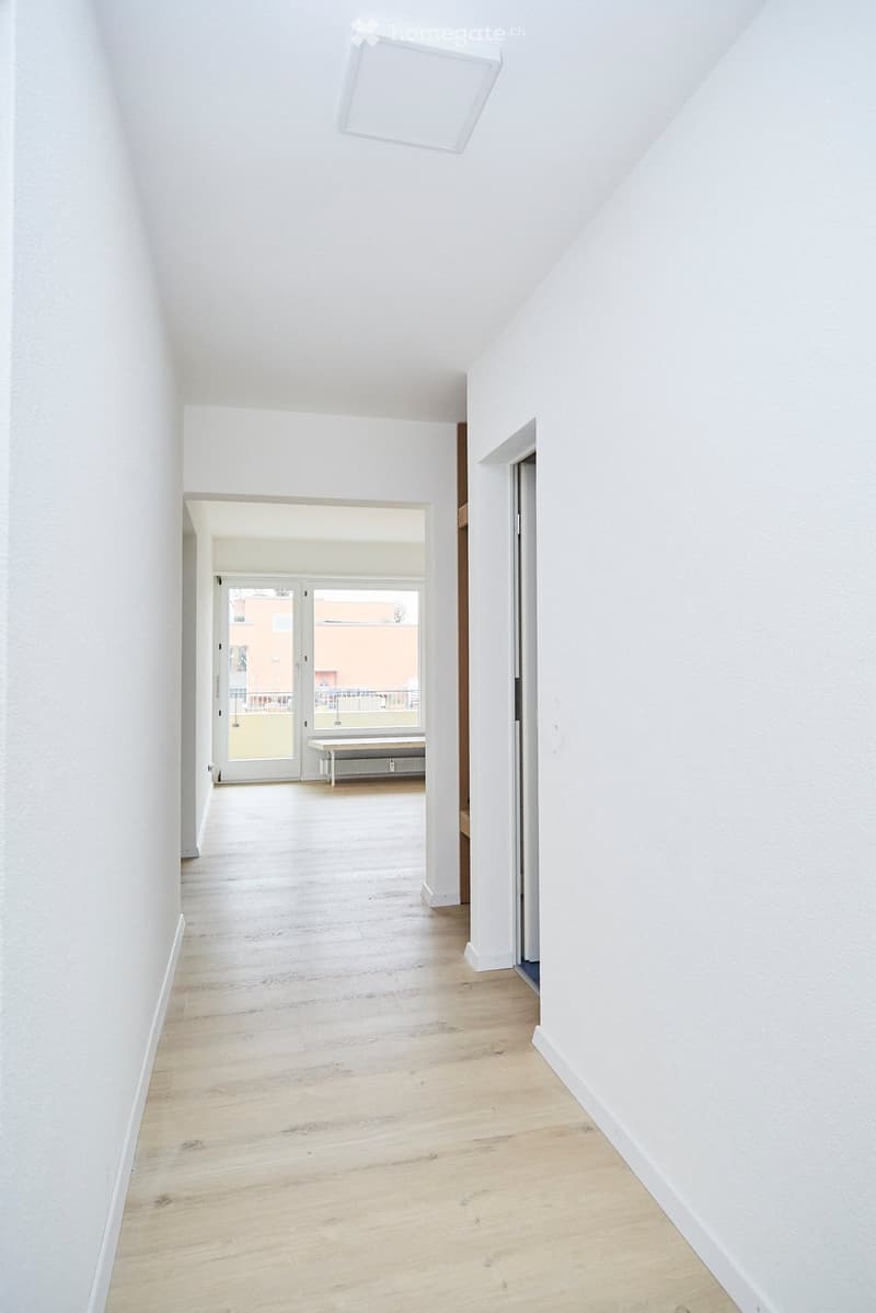 Bright, minimalist hallway with white walls, wooden flooring, and a skylight ceiling. Features an open doorway leading to a room with a bench and radiator.
