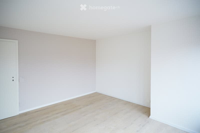 Empty room with light wooden flooring and white walls, featuring a single white door on the left side.