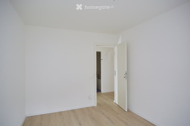 Empty room with white walls and light wooden flooring, featuring an open doorway leading to another room.