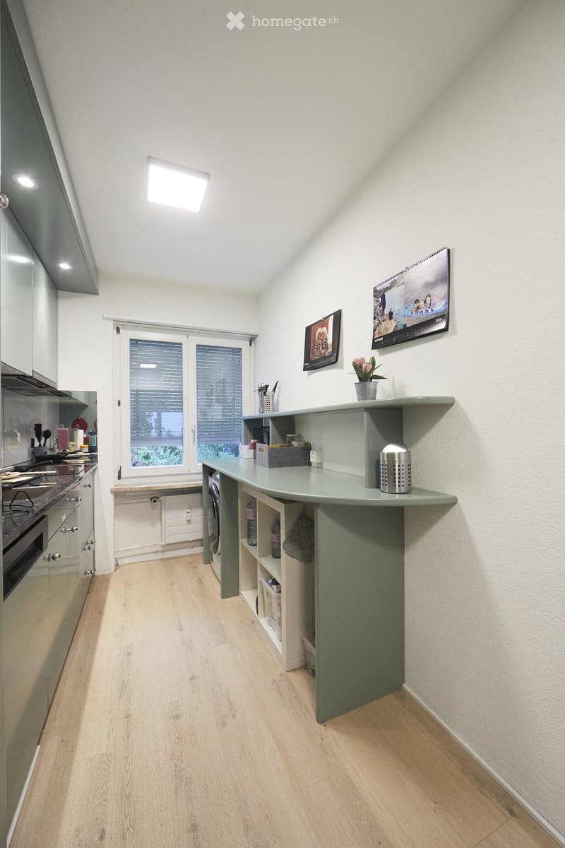 Modern kitchen with light wooden flooring, grey cabinets, and a central island counter. Features include an oven, refrigerator, and a small shelf with decorative items.