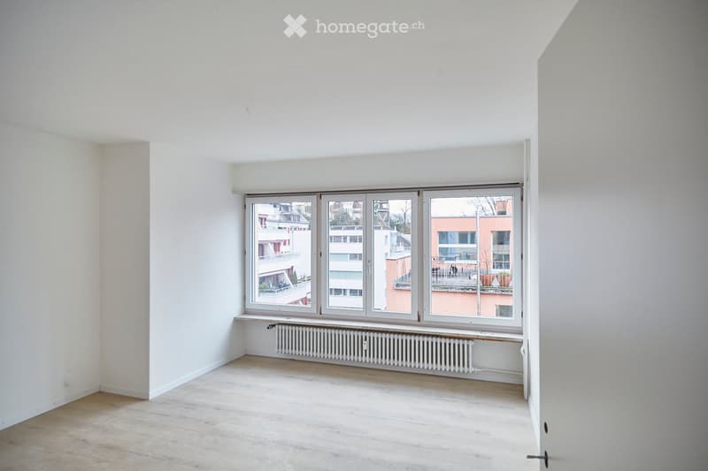 Empty room with light wooden flooring, white walls, large window offering a view of neighboring buildings, and a radiator below the window.