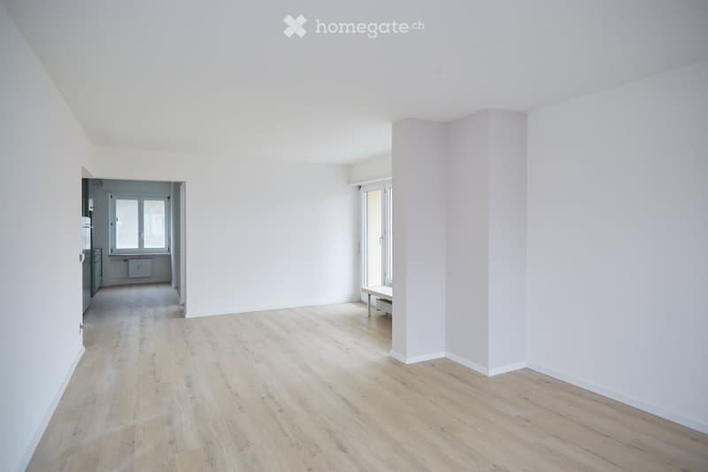 Empty room with light wooden flooring and white walls, featuring a doorway to another room and a window with a bench beneath it.