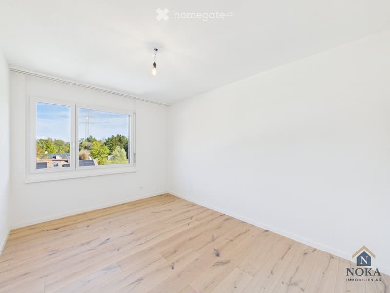 Empty room with white walls, wooden floor, and large windows offering a view of greenery outside.