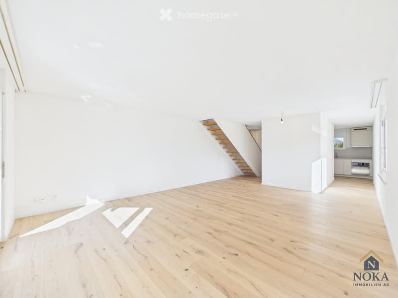 Bright, empty room with light wooden flooring and white walls. A staircase leads to an upper level, and a small kitchen area is visible in the background.