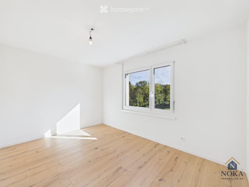 Empty room with white walls, wooden floor, and large window showing greenery outside.