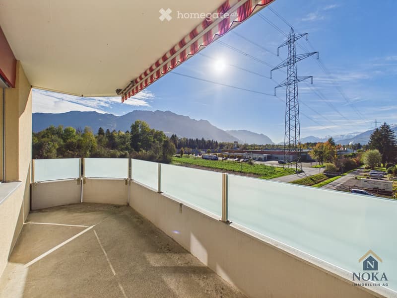 Balcony with a concrete floor, glass railing, and a striped awning, overlooking a scenic view of mountains, trees, and a power pylon.