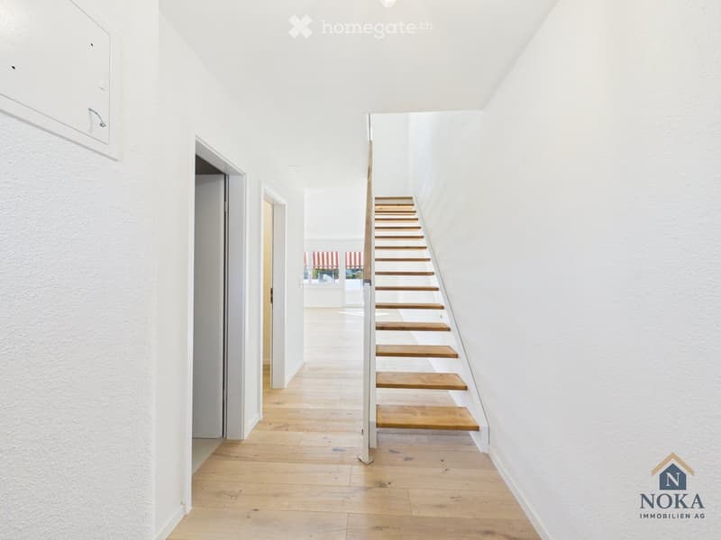 Bright hallway with wooden flooring, white walls, and a staircase leading upwards. Doors open to adjacent rooms.