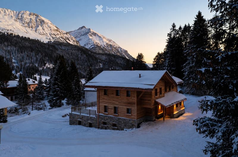 A wooden house with a stone base, snow-covered roof, and illuminated entrance, situated in a snowy mountain landscape with surrounding trees and distant peaks.