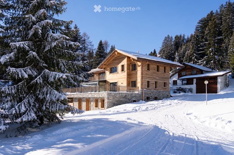 A large, modern wooden house with a stone base, multiple windows, and a balcony, surrounded by snow-covered trees and a clear blue sky.