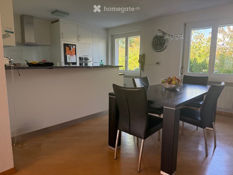 Dining room with a black dining table and chairs, a bowl of fruit, and a modern kitchen area with cabinets, a fridge, and an oven.