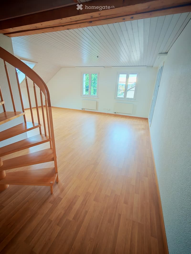 Empty room with wooden flooring, white walls, and a wooden staircase leading upwards. Windows provide natural light, and radiators are visible along the walls.