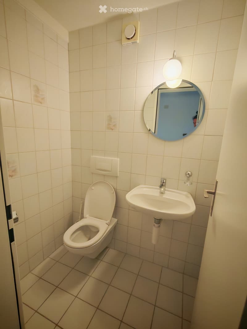 Small bathroom with white tiled walls and floor, featuring a toilet, sink, and round mirror with a built-in light.