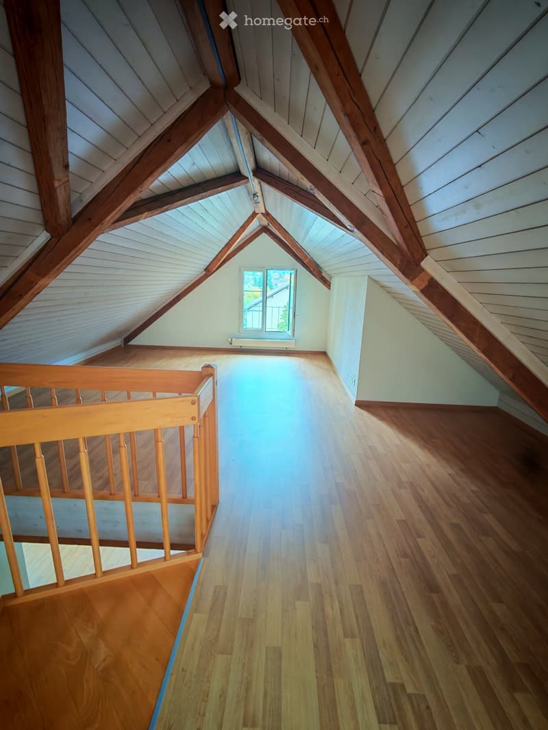 Empty attic space with wooden beams, slanted wooden ceiling, wooden floor, and a small window with a view of the outside.
