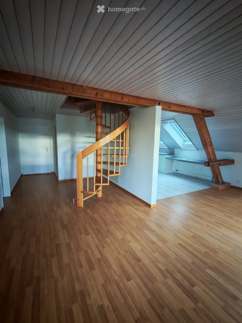 Empty room with wooden flooring, a spiral staircase, and a small kitchen area visible in the background.