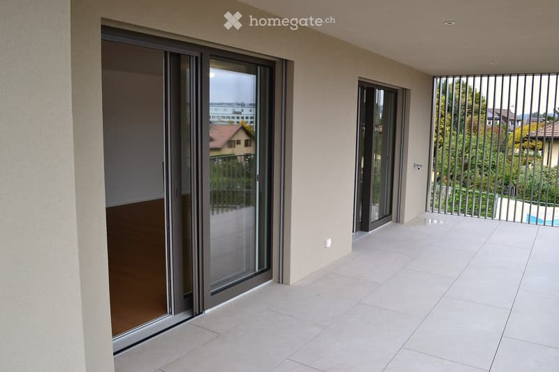 Modern balcony with large sliding glass doors, tiled floor, and metal security bars, overlooking a residential area with greenery and a swimming pool.