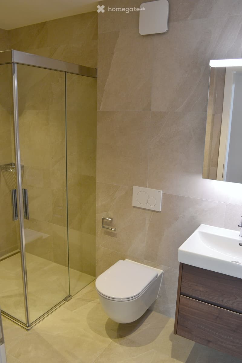 Modern bathroom with beige tiles, a white toilet, a sink with a dark wooden cabinet, and a glass shower enclosure.