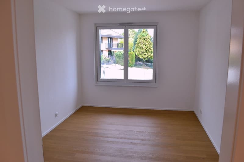 Empty room with light wooden flooring and white walls, featuring a large window with a view of a balcony and greenery outside.