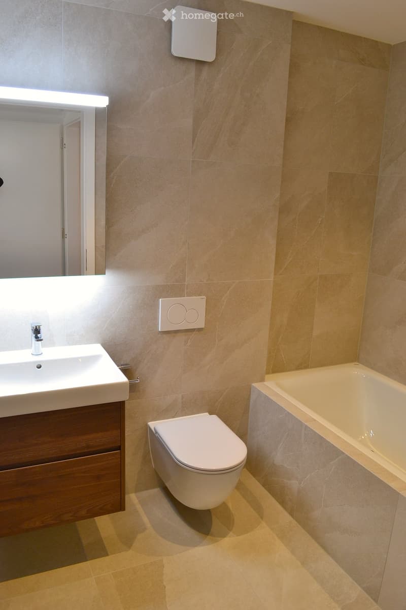 Modern bathroom with beige marble tiles, a white sink with a chrome faucet, a toilet, a bathtub, and a large mirror with built-in lighting.