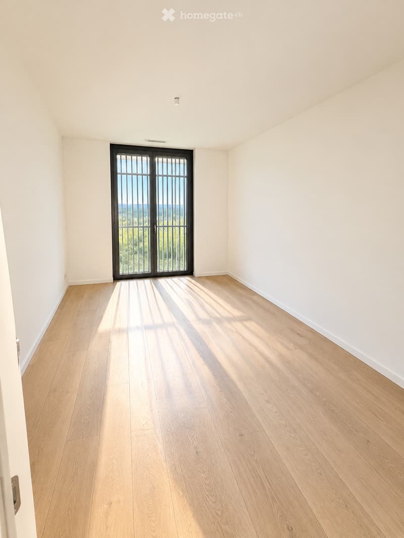 Empty room with light wooden flooring, white walls, and large windows offering a scenic view of greenery and hills.