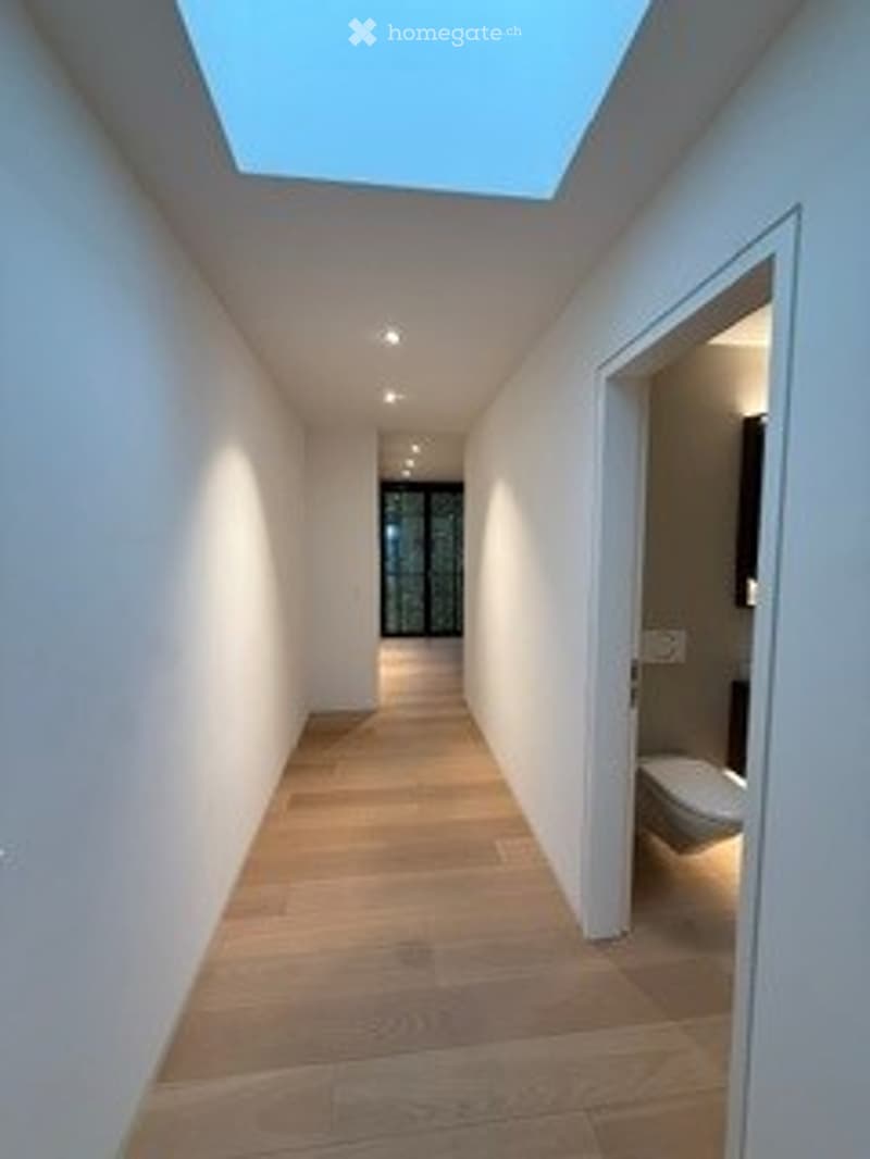 A modern, well-lit hallway with wooden flooring, white walls, and recessed ceiling lights. The hallway leads to a partially visible bathroom with a toilet and a mirror.