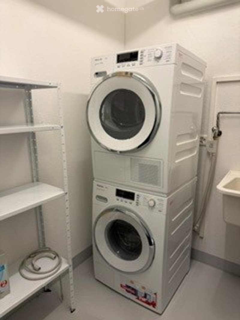Laundry area with stacked washing machine and dryer, white shelving unit, and a small sink.