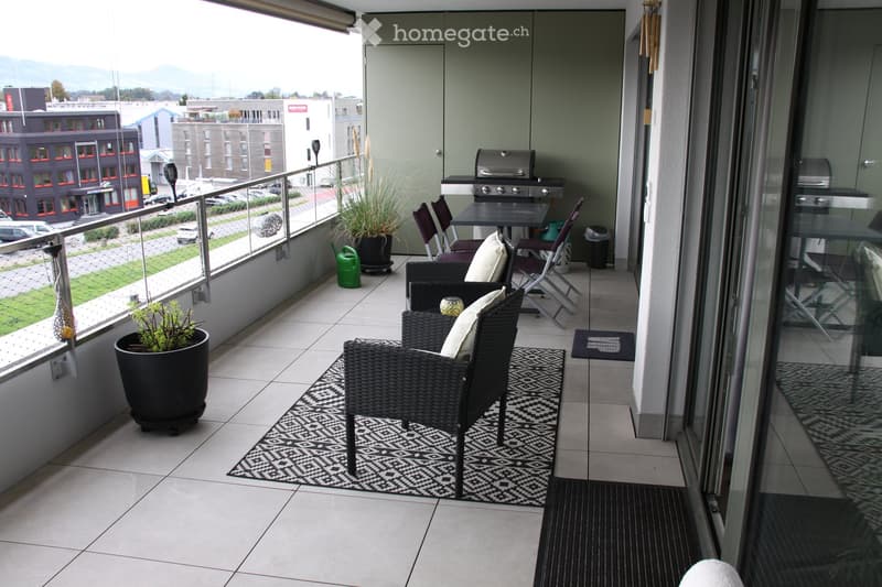 Balcony with tiled floor, black wicker chairs, small table, grill, potted plants, and rugs.