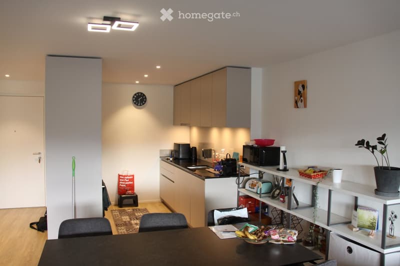 The image shows a modern, open-plan kitchen and dining area. The kitchen features light wooden cabinets, a black countertop, and various appliances including a microwave, toaster, and sink. The dining area has a black dining table with chairs, a bowl of f