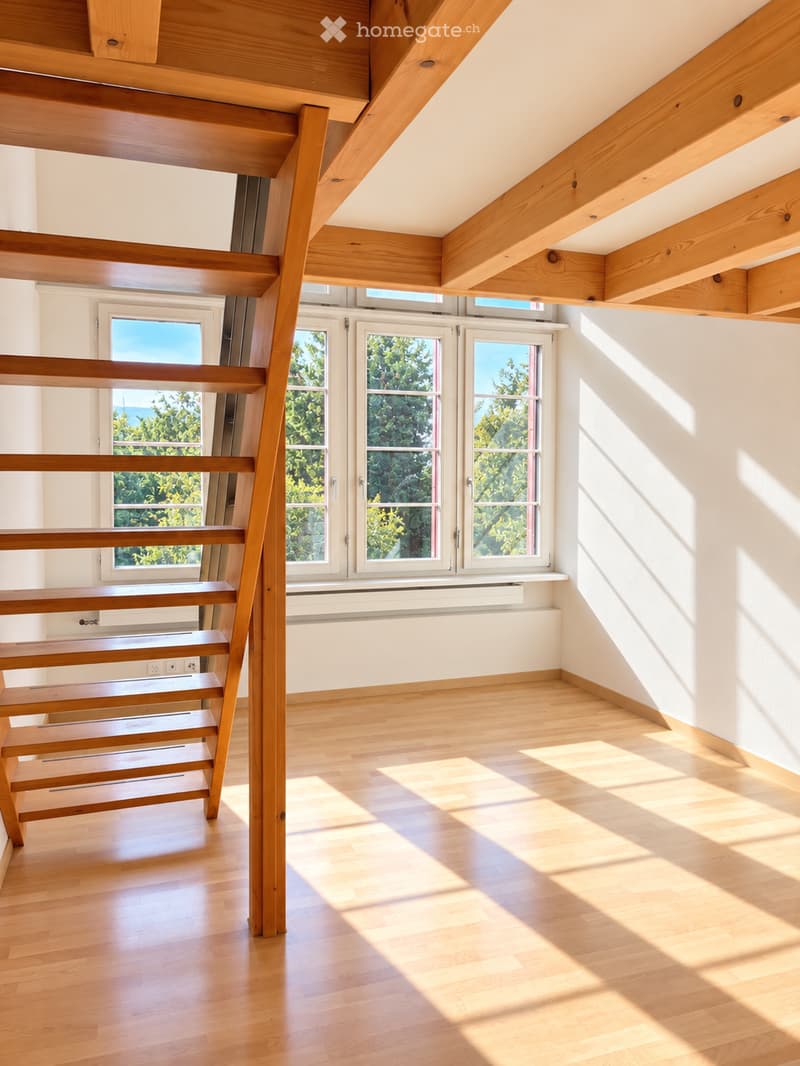 Bright, empty room with wooden flooring and exposed wooden beams on the ceiling. Large windows allow natural light to fill the space, casting shadows on the floor.