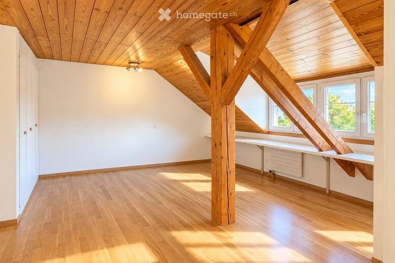 Bright, empty room with wooden floors and exposed wooden beams in the ceiling. Large windows allow natural light to fill the space. The room features built-in shelving and a small desk area.