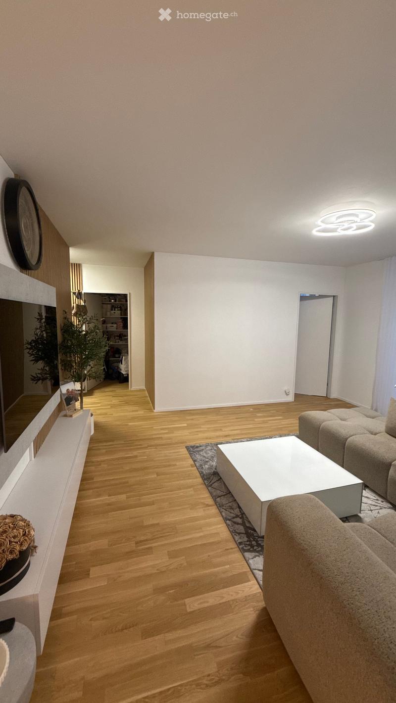 Modern living room with light wooden flooring, white walls, a grey sofa, a white coffee table, a wall-mounted TV, and a shelf with decorative items and a potted plant.