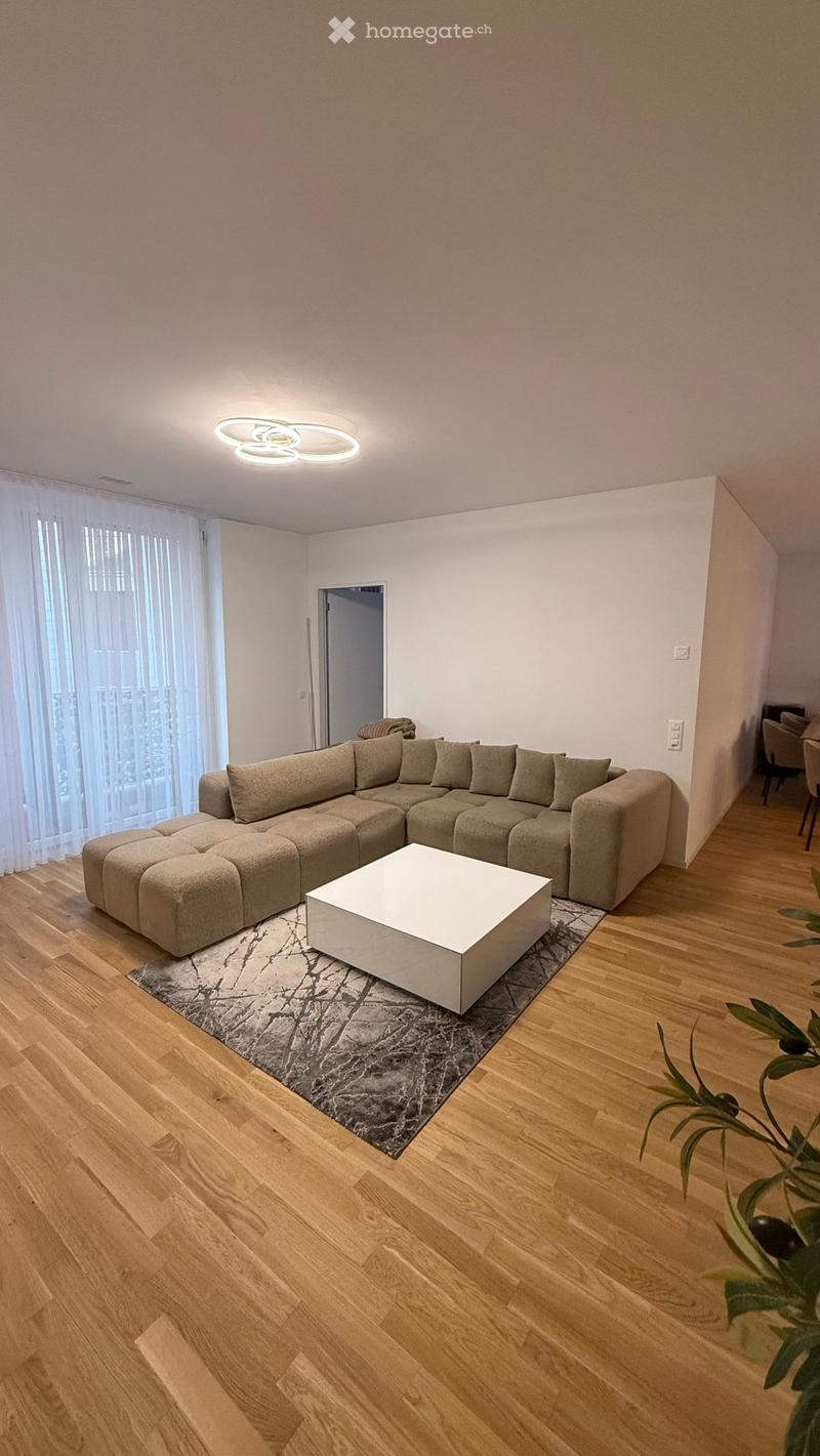 Modern living room with a large sectional sofa, white coffee table, wooden flooring, and large windows with white curtains.