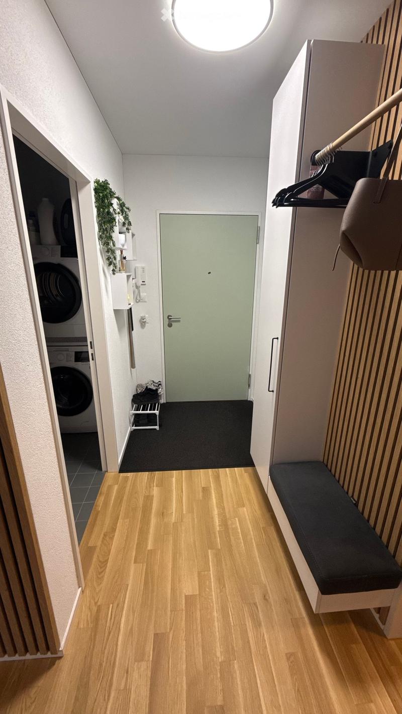 A narrow hallway with wooden flooring, a bench on the right, a door at the end, and a small closet with a hanging bag. A washing machine is visible in an adjacent room.