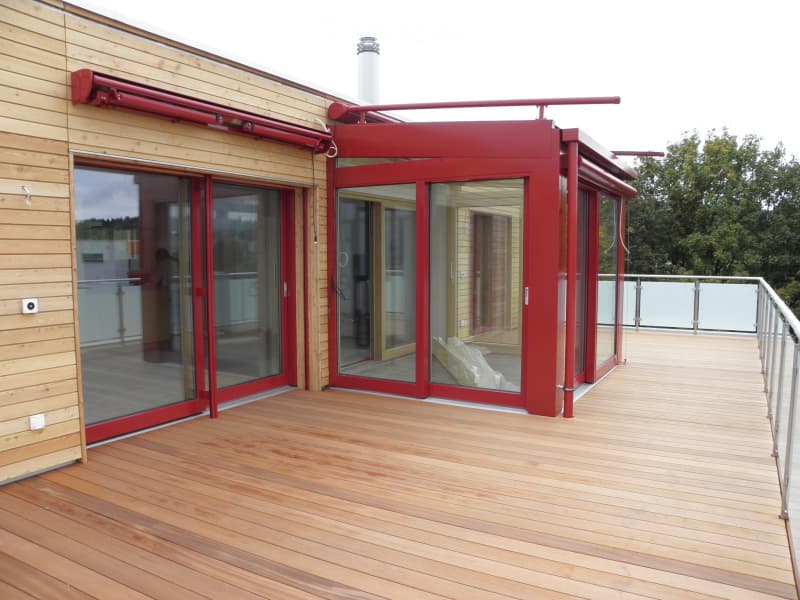 Wooden deck with red sliding glass doors leading to a covered area, featuring a metal railing and a view of greenery.