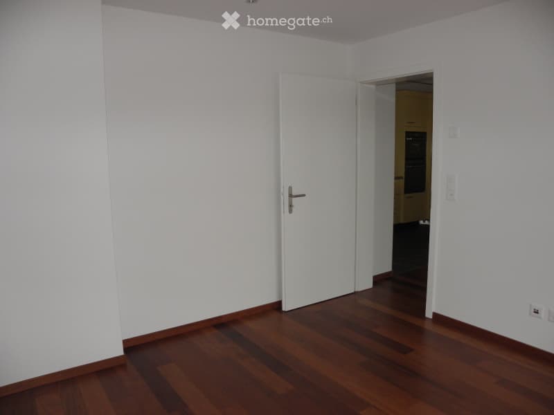 Empty room with white walls and wooden flooring, featuring an open door leading to another room.