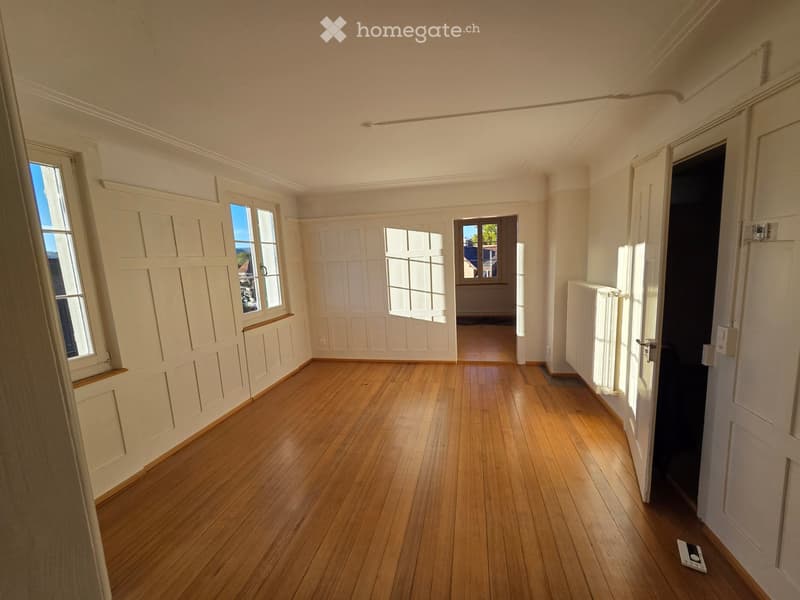 Empty room with wooden flooring, white paneled walls, and large windows allowing natural light. A radiator is visible on one wall, and a doorway leads to another room.