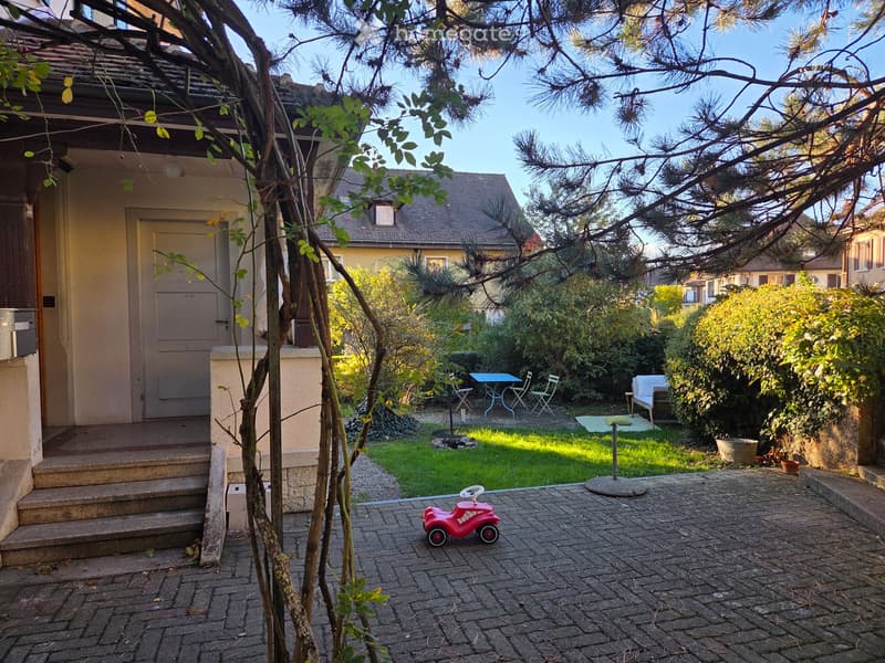Garden area with a paved patio, small lawn, outdoor furniture, and a toy car. The scene includes a house entrance with steps and a small tree in the foreground.