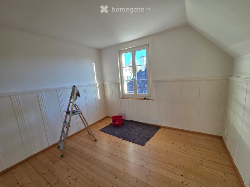 Empty room with wooden flooring, white walls, and a window. A ladder, a red bucket, and a black mat are present.